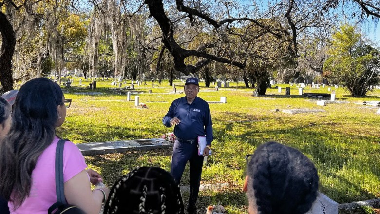 A wide exterior photograph showing historian Fred Hearns leading a group on a College Hill Black History Walking Tour in Tampa, Florida. Positioned at the center of a sunlit grassy cemetery under moss-draped oak trees, Hearns wears a navy long-sleeve collared shirt, dark pants, and a ballcap while gesturing with his hands during his presentation. In the foreground, a small tour group viewed from behind listens intently, with white gravestones scattered across the lawn behind Hearns under a bright blue sky.