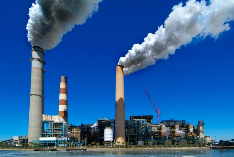 View of the Tampa Electric (TECO) power plant in Apollo Beach, featuring three tall smokestacks releasing large plumes of white exhaust into a clear blue sky, with the industrial facility at the water's edge.
