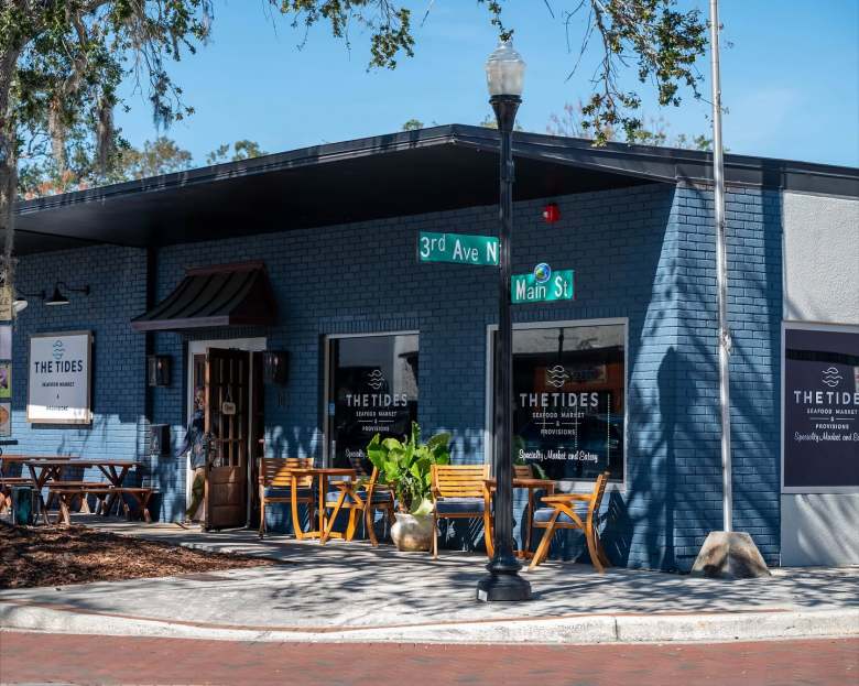 Exterior view of The Tides Seafood Market & Provisions building located at the corner of 3rd Ave N and Main St, featuring blue brick walls, large glass windows, and outdoor wooden seating.