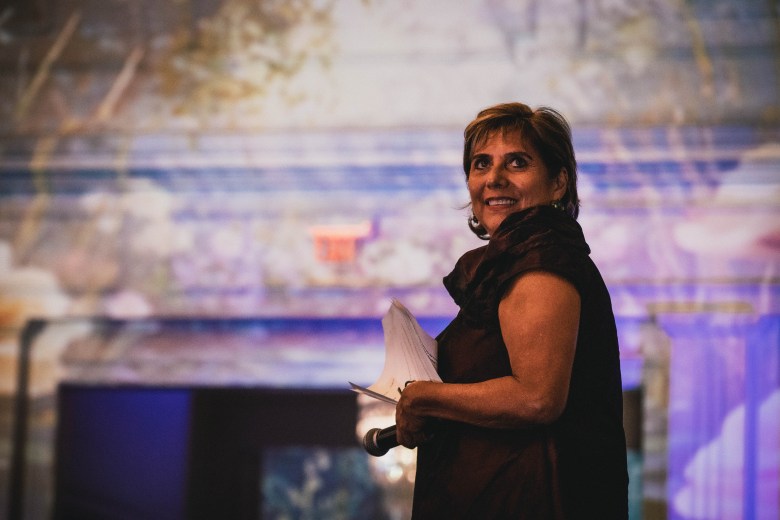 An indoor, medium close-up photograph showing founder and Artistic Director Paula Nuñez at Palma Ceia Country Club in Tampa, Florida on Sept. 18, 2026 during the Tampa City Ballet Gala & Auction. Captured from a slightly low three-quarter angle, Nuñez stands holding papers and a microphone in her hands, smiling as she looks upward and off-camera to the left. She wears a sleeveless dark brown satin top with a wide cowl neck, styled with short layered brown hair. In the softly blurred background, ambient purple and blue stage lights illuminate an event space backdrop.
