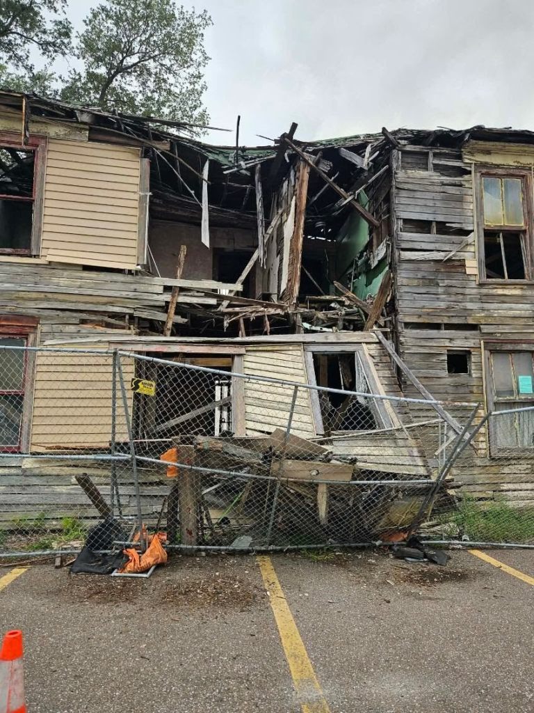 A photograph showing the severely damaged exterior structure of the historic Jackson House in Tampa, Florida on Sept. 7, 2026. The two-story historic wooden building exhibits extensive structural collapse across its central second-floor facade, exposing weathered timber framing, interior walls, and broken rafters under an overcast sky. In the foreground, a chain-link security fence stands bent and partially collapsed across an asphalt parking lot painted with yellow parking lines, next to an orange traffic cone.