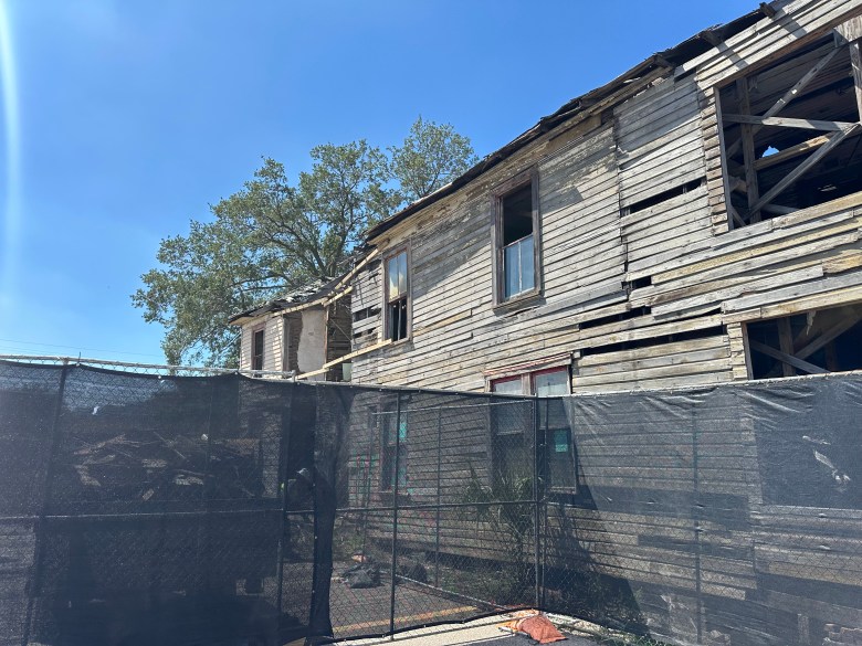 An exterior photograph by Valerie Smith showing the side profile of the historic Jackson House in Tampa, Florida on Sept. 9, 2026. Looking upward from a low angle along the weathered, unpainted wooden siding, the multi-story historic building displays missing exterior planks, exposed timber framing, and empty window frames under a clear blue sky. In the foreground, a tall black mesh construction fence obscures the lower portion of the structure along the perimeter.