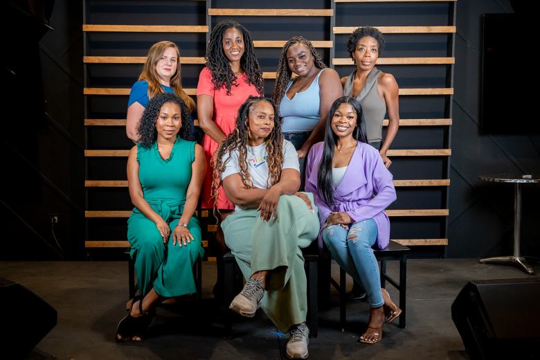 A promotional group portrait of the cast and director of Kelundra Smith's 'The Wash,' making its Florida premiere Oct. 1-11 at The Studio in St. Petersburg. Seven women pose in two rows against a dark studio background with a black-and-wood slatted wall accent. In the back row (L-R), Holly Marie Weber stands in a bright blue top, Jemier Jenkins in a pink dress with braided hair, Jada Griffin in a light blue sleeveless top, and Andresia Moseley in a grey top. Seated in the front row (L-R), Nadine Young poses in a teal sleeveless jumpsuit, director Erica Sutherlin sits centrally with crossed legs in a white top, light green trousers, and sneakers, and Marjorie Joseph sits on the right in a lilac blazer, light blue jeans, and heels.