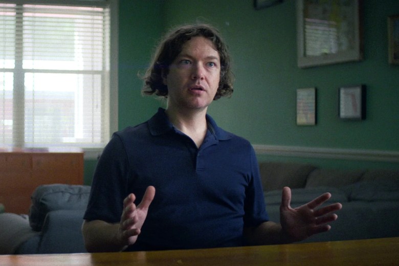 A medium close-up documentary screengrab showing media consultant Tim Burke in 'Untold: The Girlfriend Who Didn't Exist'. Centered at a wooden table, Burke wears a short-sleeved dark navy polo shirt and has shoulder-length, wavy brown hair, gesturing with open hands while speaking during an interview. Behind him, a softly lit room features sage green walls, framed artwork, a sofa, and a window with white blinds on the left.