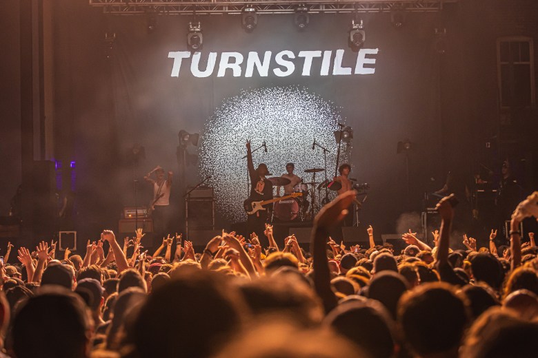 A wide concert performance photograph by Dave Decker showing hardcore punk band Turnstile playing at Jannus Live in St. Petersburg, Florida on Oct. 14, 2022. Captured from behind a dense, packed crowd of fans with their arms raised in the air, the band performs on a brightly lit stage under a prominent white backdrop displaying the word "TURNSTILE" in bold italicized lettering above a circular, patterned lighting fixture.