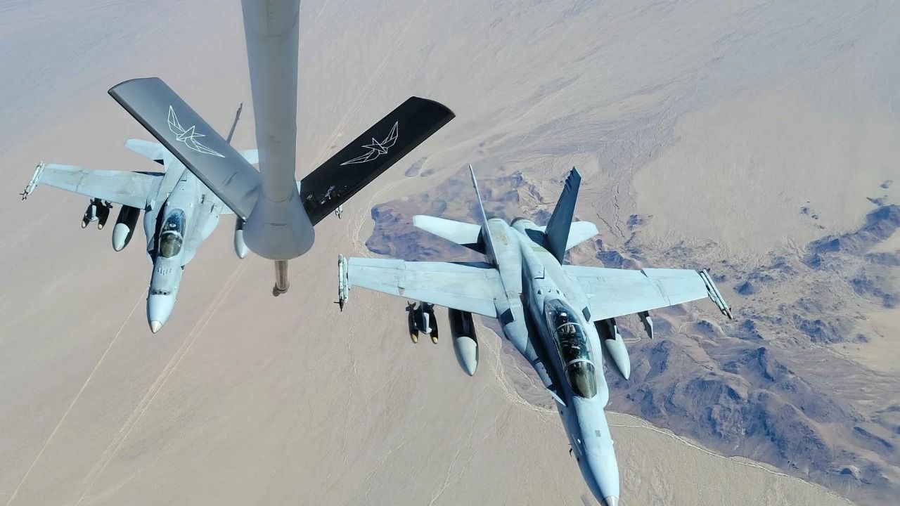 Two military aircraft fly alongside an aerial refueling tanker over a desert landscape.