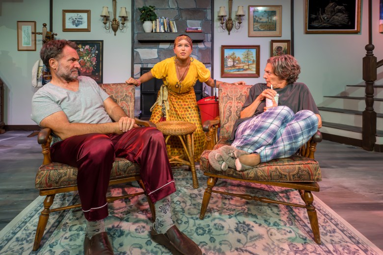 A stage production photograph by Chaz D. Photography showing (L-R) Michael Gregory, Kayah Bonilla-Mas and Jill Schroeder in 'Vanya and Sonya and Masha and Spike' at Off-Central Players in St. Petersburg, Florida. Set inside a cozy living room, Michael Gregory sits on the left in a wooden armchair wearing a grey t-shirt, burgundy satin pajama pants, and slippers, facing Jill Schroeder on the right, who lounges in a matching armchair in blue plaid pajama pants and a grey top while sipping from a cup with a straw. In the center background, Kayah Bonilla-Mas stands between them in a yellow patterned dress with a red headscarf, leaning forward over a small round woven table against a backdrop framed by framed artwork, wall sconces, and a staircase.