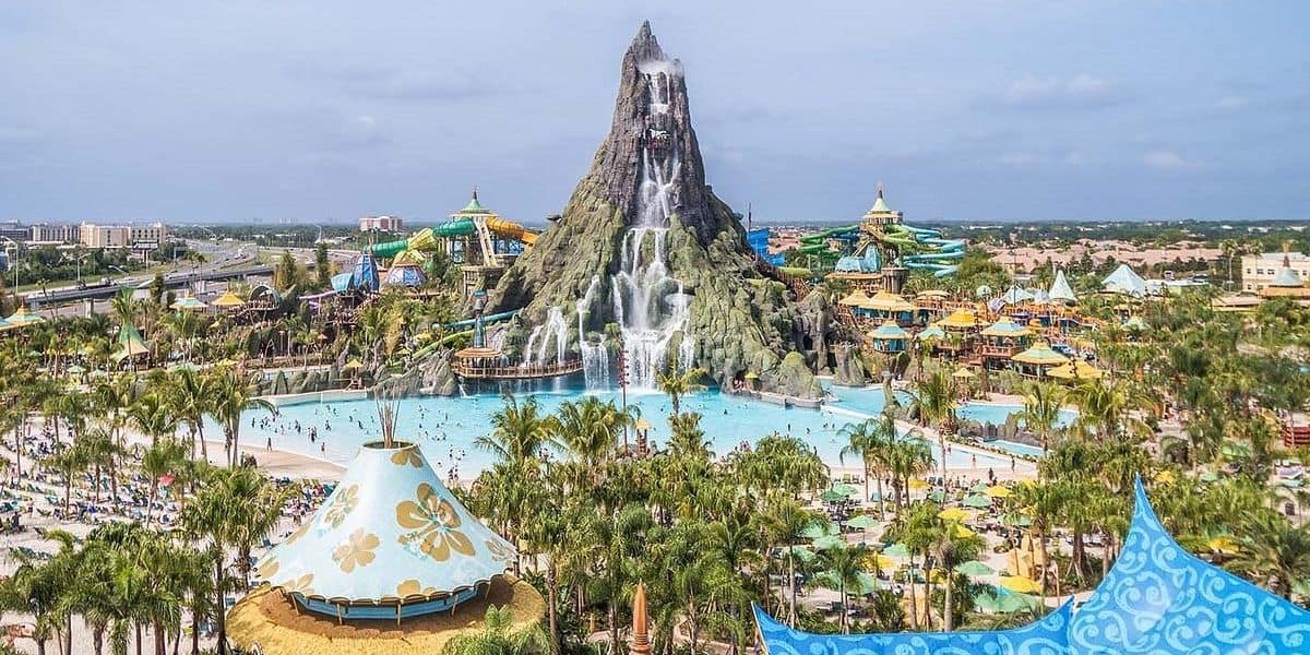 Aerial view of Volcano Bay water park at Universal Orlando Resort featuring a grand, volcano-like structure with cascading waterfalls. Surrounding the volcano are pools, slides, and lush greenery. Visitors enjoy the sunny day among the attractions and palm trees, easily navigating with the virtual line system.