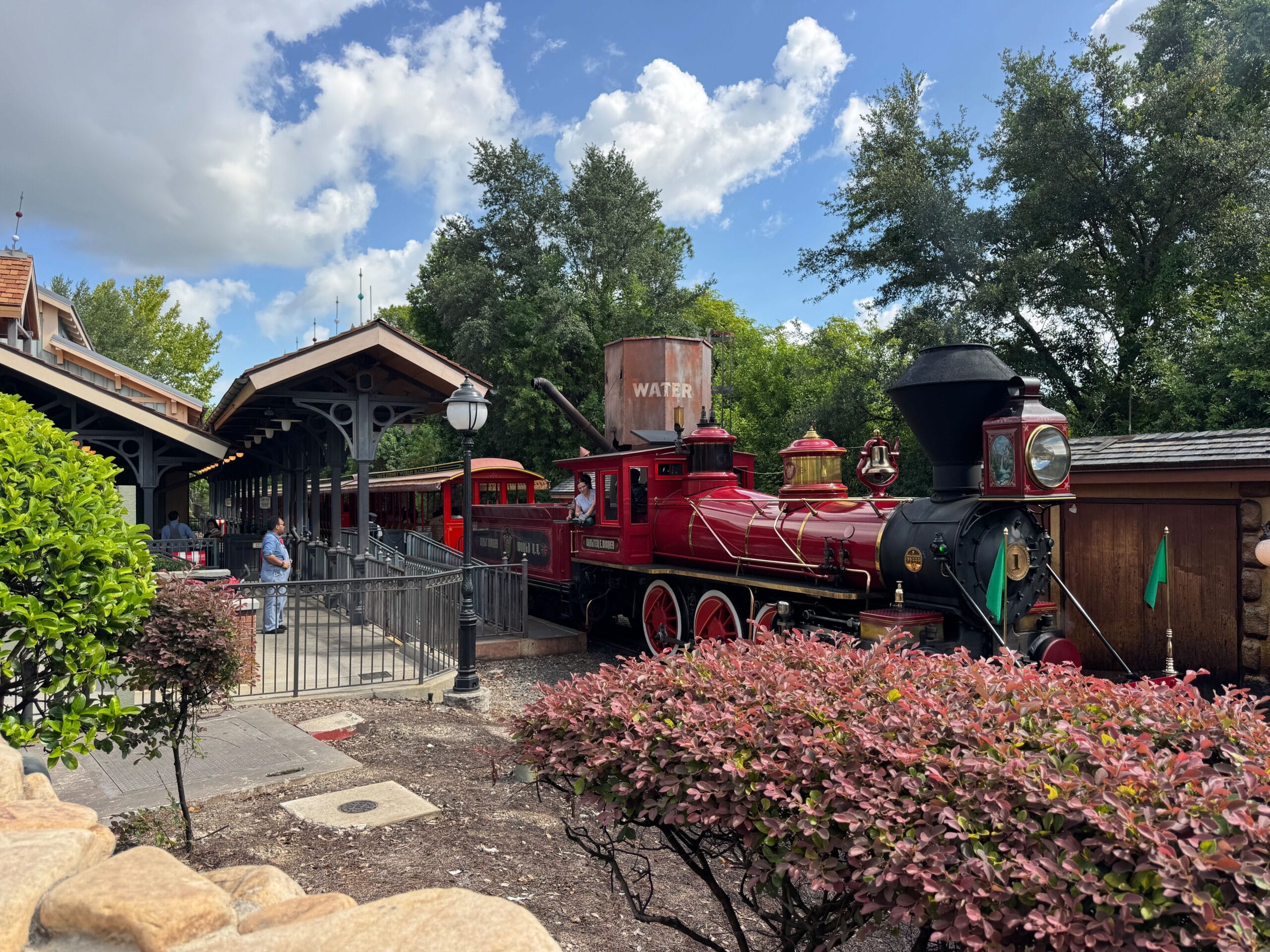 A bright red vintage steam train, like the Walt Disney World Railroad, waits at a station amid trees and greenery.