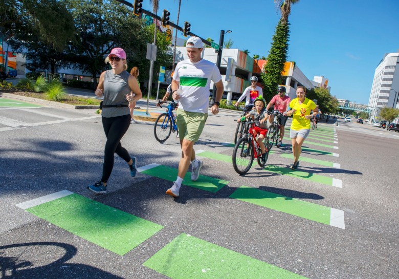 A sunny, action-oriented outdoor photograph showing participants during Walk Bike Tampa World Car-Free Day on Sept. 22, 2024. Captured from a low angle, a group of pedestrians and cyclists cross a wide street along bright green painted crosswalk markers under a clear blue sky. In the foreground, a woman wearing a pink baseball cap, grey tank top, black leggings, and sneakers runs alongside a man wearing a white backwards cap, a white shirt with a green stripe, tan shorts, and running shoes. Behind them, several adults and a child on bicycles, as well as a runner in a yellow t-shirt, travel across the intersection framed by palm trees, overhead traffic lights, and city buildings.