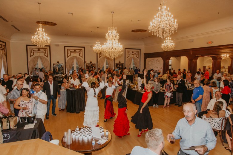 A wide indoor photograph of flamenco dancers performing in the center ballroom during the Wines of Spain event at the Centro Asturiano de Tampa in Ybor City, Florida on Oct. 16, 2024. Under grand crystal chandeliers suspended from high ceilings, dancers in vibrant red and white ruffled flamenco skirts twirl in the middle of a hardwood dance floor. Surrounding the performance floor, guests socialize around tall tasting tables and wine display stations stocked with glasses, bottles, and appetizers.