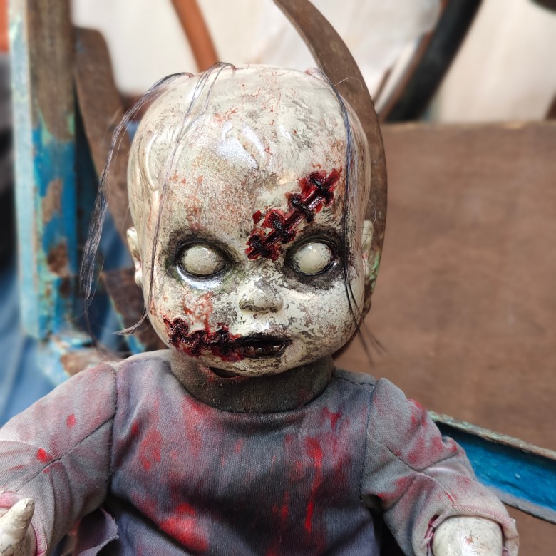 A close-up horror photograph of a weathered, blood-stained porcelain doll displayed as a Halloween prop or haunted house decoration. The doll features pale, cracked white skin, milky white blank eyes, a stitched wound across its forehead, dark red fake blood smeared around its mouth and torso, thin wisps of dark hair, and a worn grey shirt, set against a rustic metal and wood background.