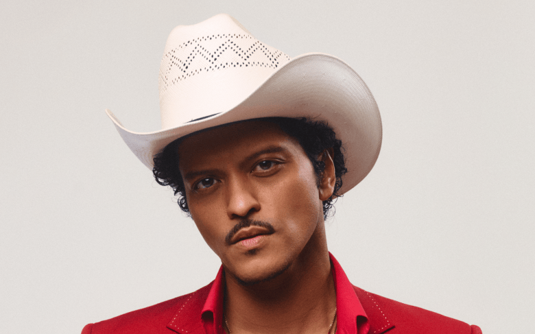 A close-up promotional portrait photograph of singer-songwriter Bruno Mars wearing a white cowboy hat. Tilting his head slightly toward his right, Mars looks directly forward with a subtle mustache, dark eyes, and short curly hair visible under the brim of a cream-colored perforated western hat. He is dressed in a vibrant red collared jacket open at the neck, set against a plain, neutral off-white studio background.