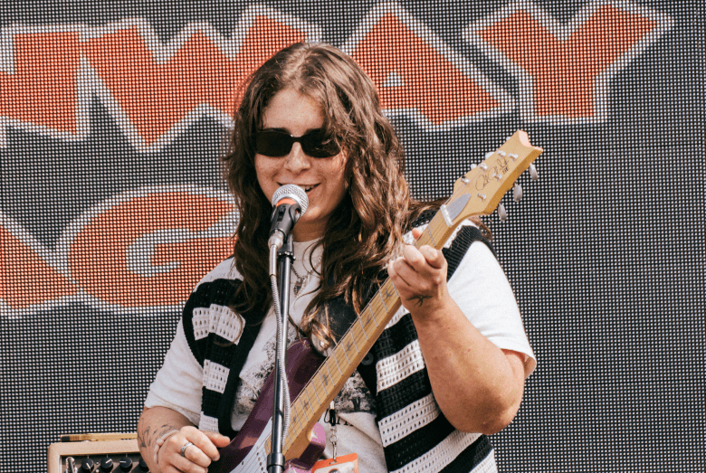 A high-energy concert photograph showing the guitarist and vocalist of indie rock band Little Giver performing at Gasparilla Music Festival at Meridian Fields in Tampa, Florida on April 10, 2026. Captured mid-performance, the musician wears dark sunglasses, a white graphic t-shirt, and a black-and-white patterned vest while playing a sunburst electric guitar and singing into a stage microphone. Set against a dark digital LED backdrop displaying red pixelated text, the artist plays on stage under bright daylight.