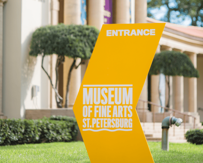 A close-up outdoor photograph of a bright yellow entrance sign standing on a green lawn in front of the Museum of Fine Arts St. Petersburg in St. Petersburg, Florida on Oct. 14, 2019. Credit to City of St. Pete via Flickr, the sharp foreground shot showcases an angled yellow directional sign reading "ENTRANCE" at the top in white text above the official white "MUSEUM OF FINE ARTS ST.PETERSBURG" logo. In the soft-focus background, manicured green hedges, a pipe fitting, decorative trees, and the museum's classical stone columns stand under sunny daylight.