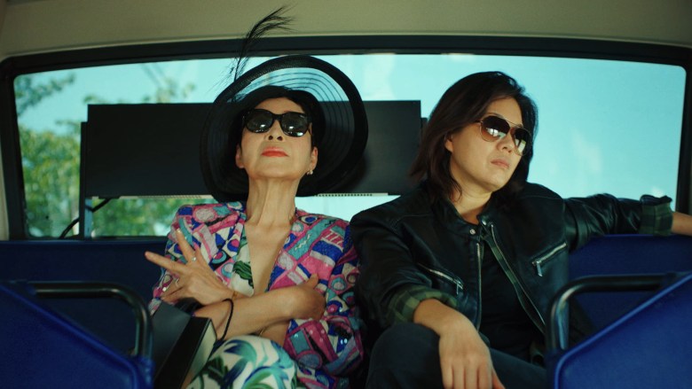 A film production still from "Manok." Captured inside a open-air vehicle against a bright blue sky, two figures sit side by side on blue seats wearing sunglasses. On the left, a person reclines backward with their head tilted up, wearing a wide-brimmed black hat with a feather accent and a vibrant, multicolored geometric jacket over floral patterned trousers while holding a peace sign. On the right, a person with shoulder-length dark hair rests their arm along the seat back, wearing a black leather jacket, dark top, and jeans while looking toward the right.