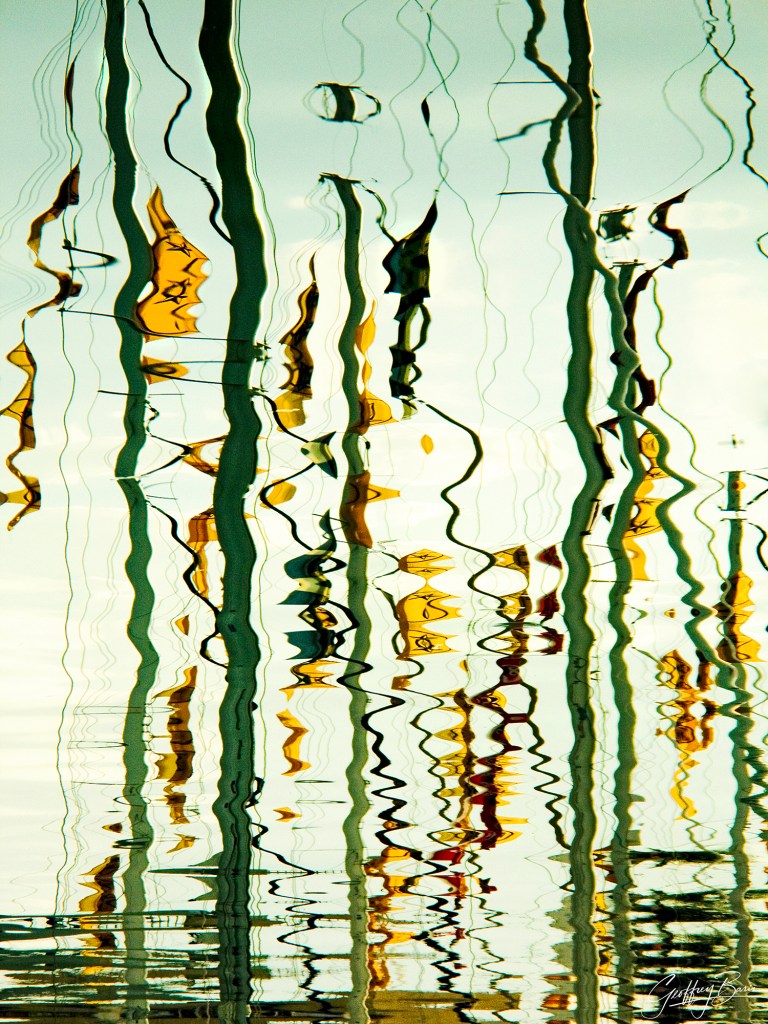 An abstract color fine art photograph titled "Wired" by photographer Geoffrey Baris (rear printed to plexiglass) depicting water reflections. Captured in a vertical framing set within a dark grey mat border, the image presents distorted, wavy reflections of sailboat masts, rigging lines, and yellow and dark nautical flags shimmering across gentle water ripples against a bright, pale sky-colored background. The artist's signature, "Geoffrey Baris," appears in white script in the lower right corner.