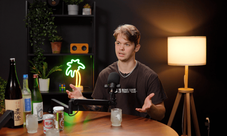 An indoor studio photograph showing podcast guest Noah Sain at the CL TV studio in Tampa, Florida on Sept. 17, 2026. Seated at a round wooden table, Sain wears a dark short-sleeved t-shirt and silver chain necklace while gesturing open-handed with both hands as he speaks into a studio microphone. Set in front of him on the table are several bottles of sake, a glass, and a small can. In the background, a green neon palm tree light glows on a dark shelf unit on the left, next to a illuminated tripod floor lamp on the right under warm, low studio lighting.