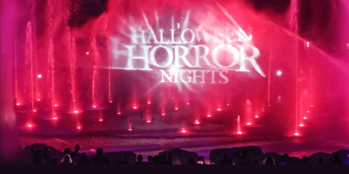 Bright pink and red water fountains glow at night as "Halloween Horror Nights 2025" is projected onto the mist above. Silhouetted fans gather, drawn to this fan-favorite spectacle in the illuminated evening at Universal Orlando Resort.