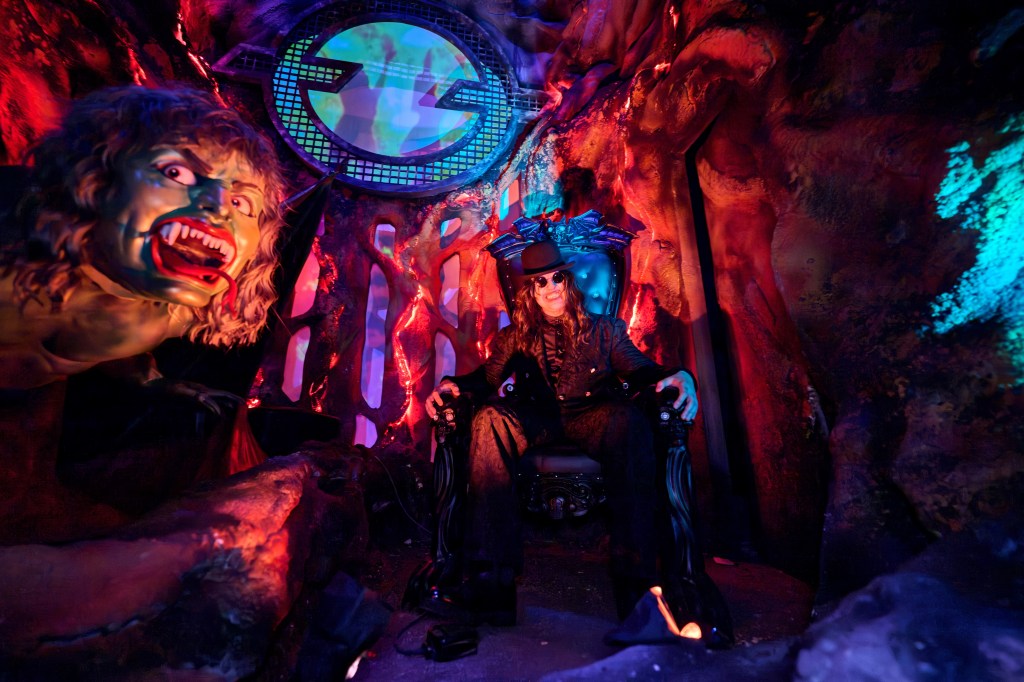 Ozzy Osbourne sits on a throne in a dark, Halloween-themed room with a monster figure to his left.