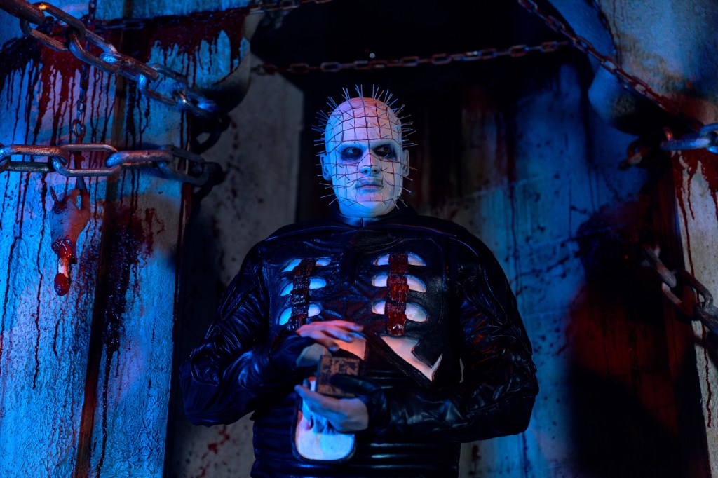 Pinhead from Hellraiser, in a dark, bloody room with chains, holding a puzzle box.
