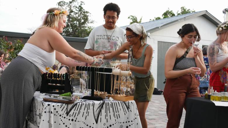 A candid outdoor photograph showing visitors exploring an artisan vendor market during Heightened Senses in Tampa, Florida. Set in a paved outdoor courtyard on an overcast evening, three women wearing tank tops, skirts, and flower crowns browse handmade jewelry display stands covered in necklaces alongside a man in a grey t-shirt, while a black table setup with bottled goods is visible on the right.
