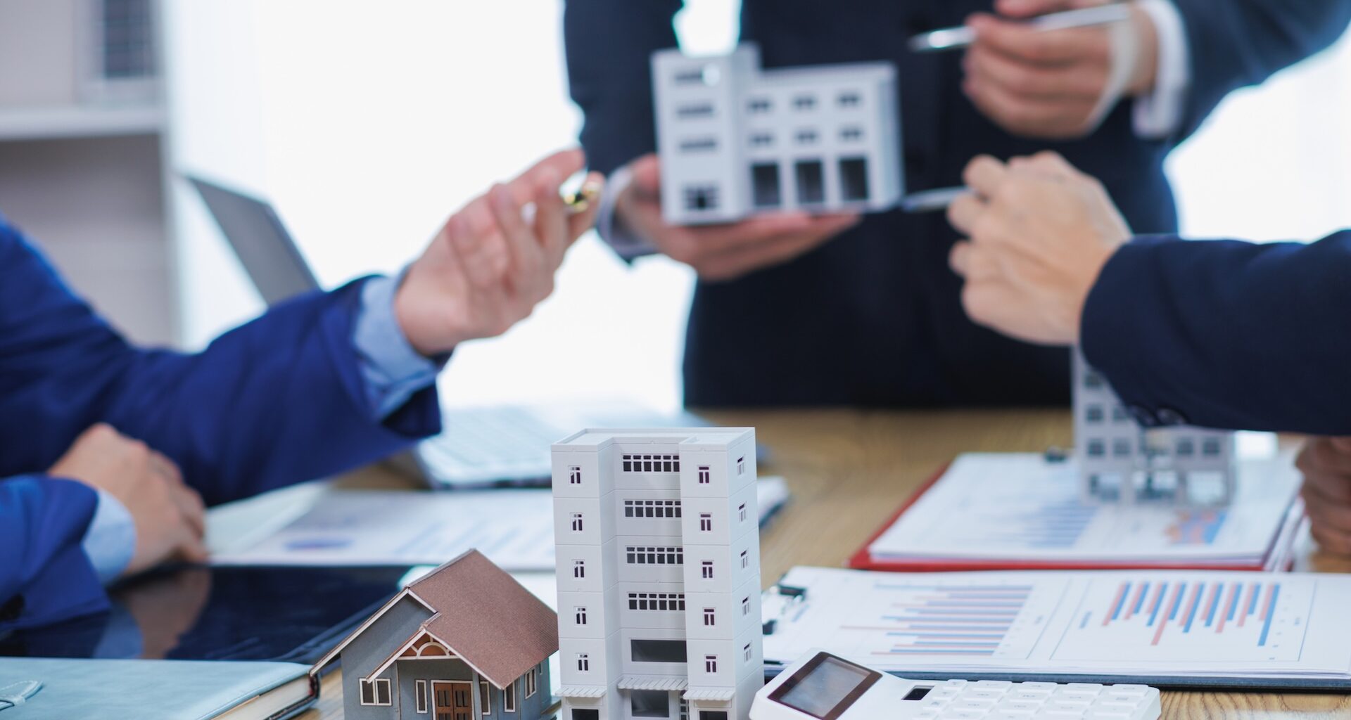 Three people discuss real estate using mini models of buildings on a table with charts and a calculator.