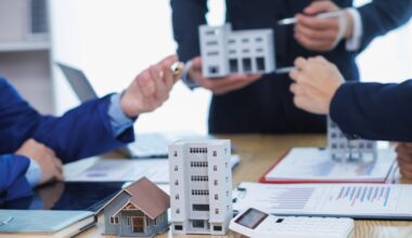 Three people discuss real estate using mini models of buildings on a table with charts and a calculator.