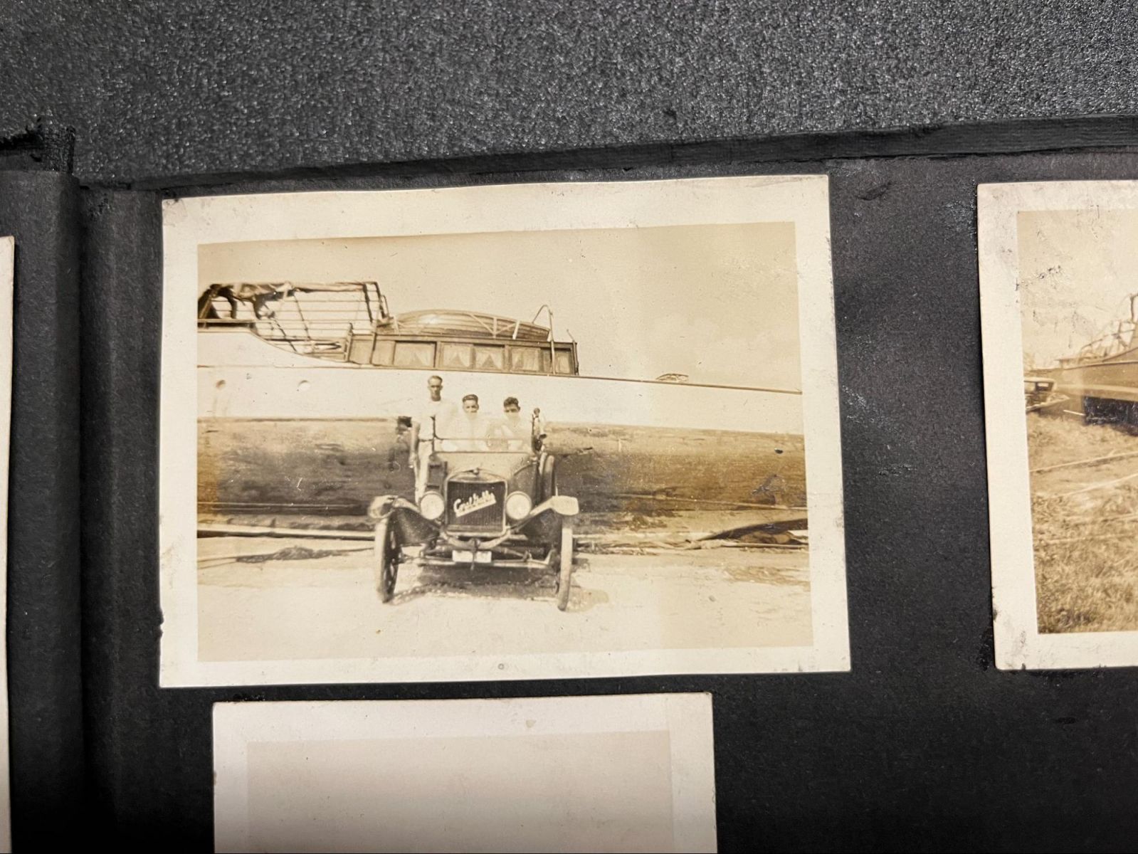 Sepia photo of people in a car parked next to a large boat on land, leaning to one side.