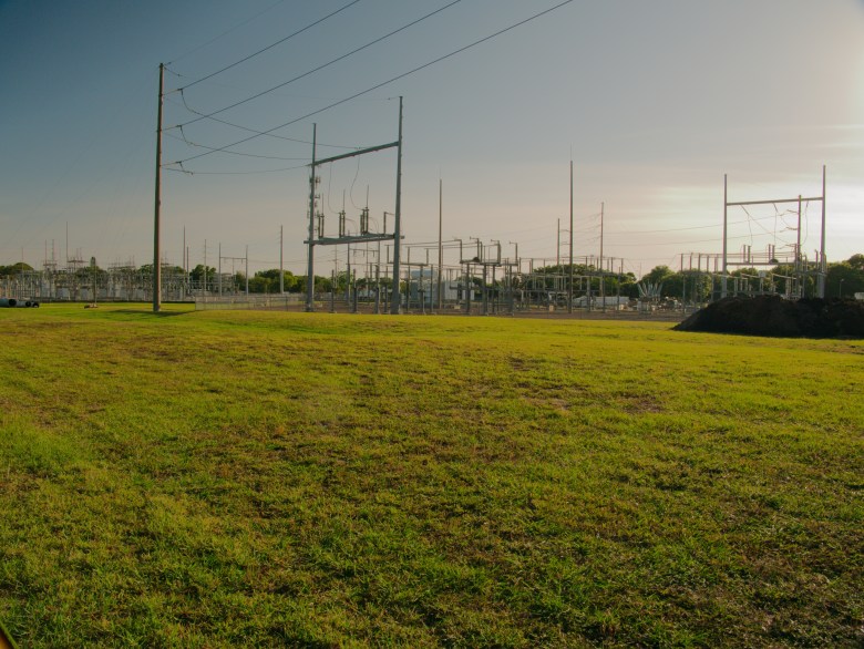 A wide landscape photograph by Del Harper showing an outdoor electric power substation set in a grassy field near sunset in St. Petersburg, Florida. Captured from across an expansive green lawn in the foreground, the image features tall wooden utility poles, steel support frames, high-voltage power lines, and electrical transformers stretching across the horizon under a hazy blue and warm yellow-tinted sky.