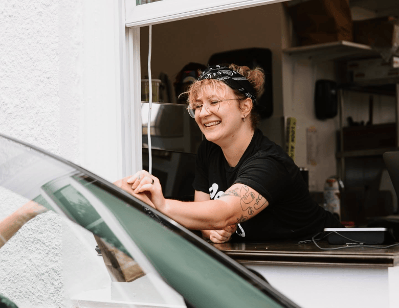 A candid exterior photograph showing a staff member working at the Pete's General drive-thru location in St. Petersburg, Florida. Leaning forward through a white-framed service window, a smiling worker with blonde hair tied in a bandana and wearing glasses, a black t-shirt, and arm tattoos hands an order to a driver inside a vehicle. The close-up shot captures the interaction from outside the car, with the white exterior wall of the building frame on the left and the interior shop counter in the background.