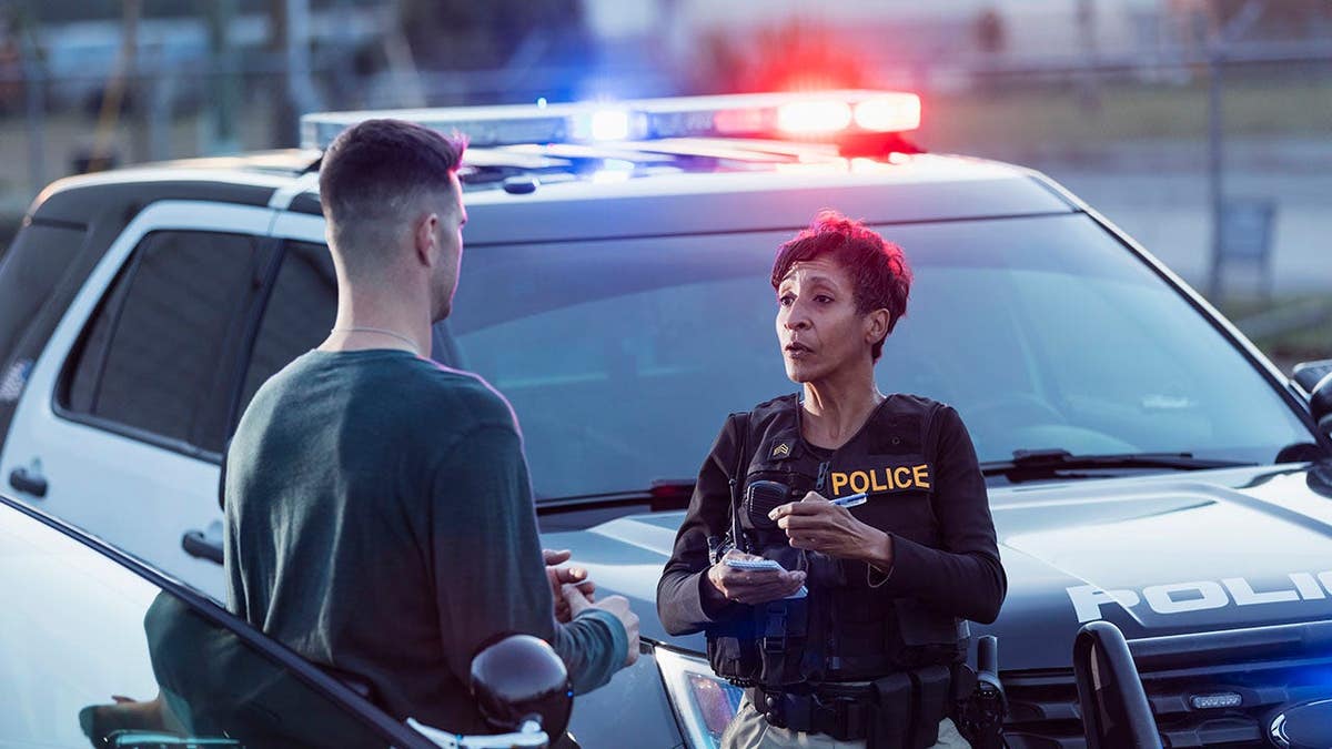 A policewoman taking a statement from a civilian outside her patrol car. The officer is a mature African-American woman in her 40s. She is talking with a young man in his 20s.