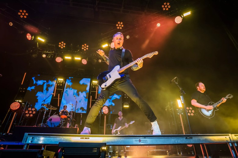 A wide-angle live performance photograph showing pop-punk band Yellowcard performing on stage at Yuengling Center in Tampa, Florida on July 23, 2023. Captured from low in front of the stage, frontman Ryan Key leaps into the air with his legs spread wide atop a metal stage platform while playing a white electric guitar under bright yellow and orange spotlights. Behind him, bandmates perform on bass, guitar, and drums set against a backdrop illuminated with blue sky and cloud visuals.