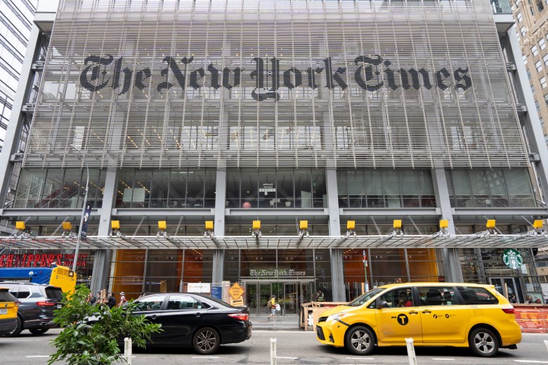 Exterior view of The New York Times Building entrance on 8th Avenue in Midtown Manhattan with a yellow taxi driving past.