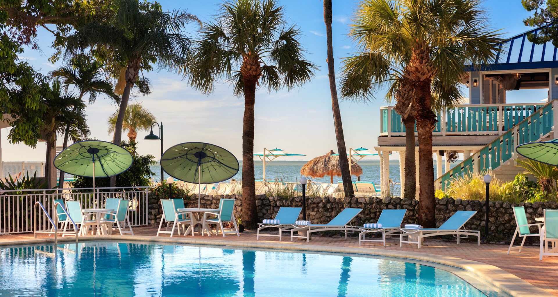 An outdoor pool area along the beach at SkyBeach Resort in St. Pete, featuring blue lounge chairs, white tables with green umbrellas, and palm trees on a sunny day.