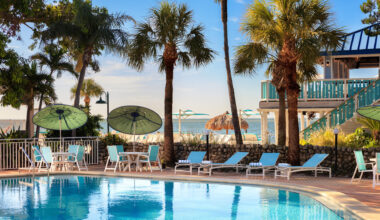 An outdoor pool area along the beach at SkyBeach Resort in St. Pete, featuring blue lounge chairs, white tables with green umbrellas, and palm trees on a sunny day.