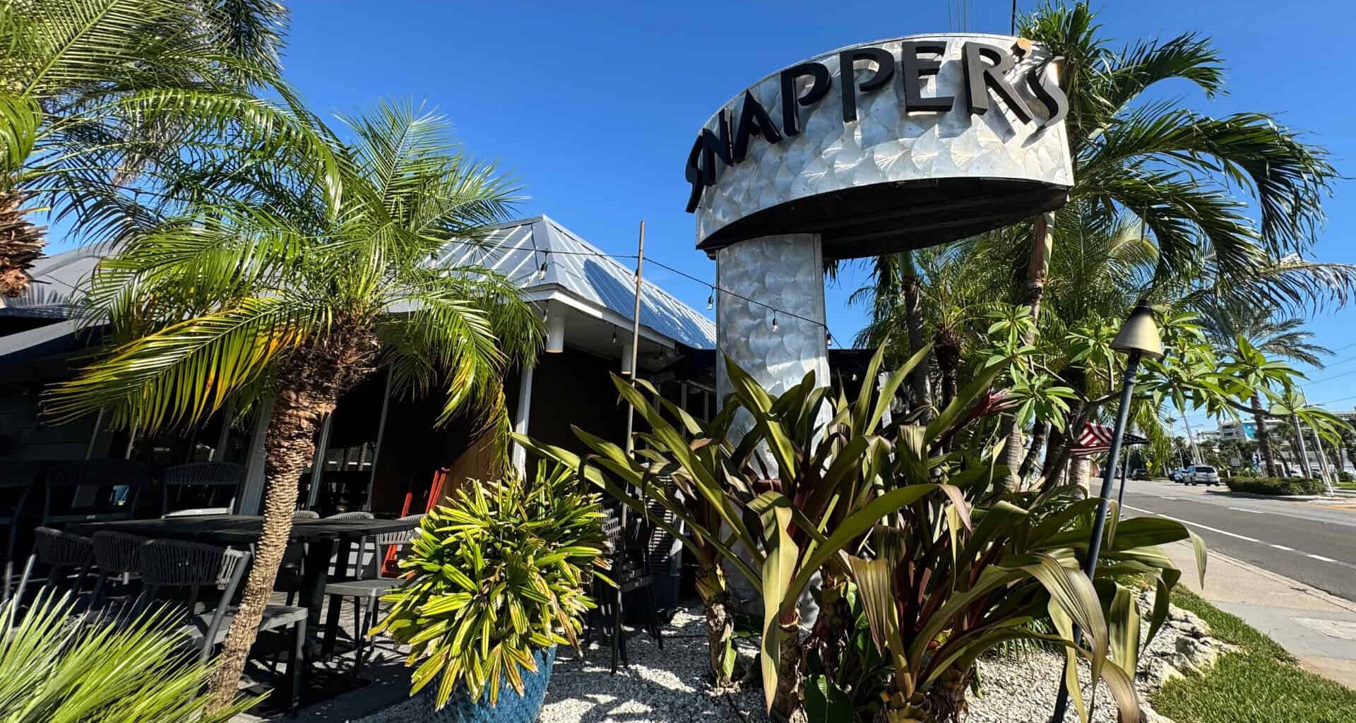 The outside seating area for Snappers Sea Grill, filled with tropical plants and palm trees under a clear blue sky.