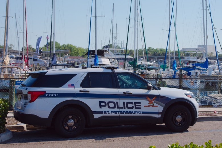 A side-profile photograph by Del Harper showing a white St. Petersburg Police vehicle at the municipal marina in St. Petersburg, Florida on March 28, 2026. The Ford Utility cruiser, displaying blue "POLICE" and "ST. PETERSBURG" side door graphics, unit number 2322, and an American flag decal on the rear quarter panel, is parked outdoors in bright daylight. In the background, tall sailboat masts and rigged boats fill the marina docks under a clear blue sky.