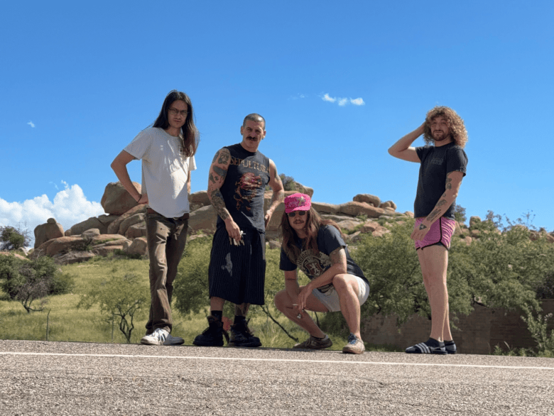 An outdoor group portrait photograph showing the band starsdontmeananything posing along the edge of a paved road against a desert landscape. Four band members stand and crouch outdoors under a clear blue sky with rocky hills and sparse green trees in the background. On the far left, a band member with long brown hair stands wearing a white t-shirt, brown trousers, and white sneakers. To their right, a band member with visible arm tattoos stands wearing a dark sleeveless graphic shirt, long dark shorts, and dark boots. Crouched in the middle, a third member wearing a pink cap, dark t-shirt, and light shorts looks toward the camera. On the far right, a fourth member with curly hair stands in profile wearing a dark shirt and pink shorts, touching their hair under bright daylight.
