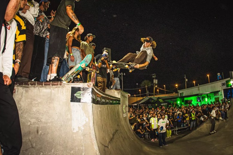 A high-action night photograph showing a skateboarder launching air mid-trick during the Tampa Am concrete jam at Skatepark of Tampa in Tampa, Florida on Oct. 18, 2025. Captured mid-flight above a concrete bowl ramp, the skater grabs their board while airborne against a dark night sky. Spectators line the top platform edge of the ramp on the left, while a large crowd watches from the lower viewing area surrounding the lighted concrete course in the background.