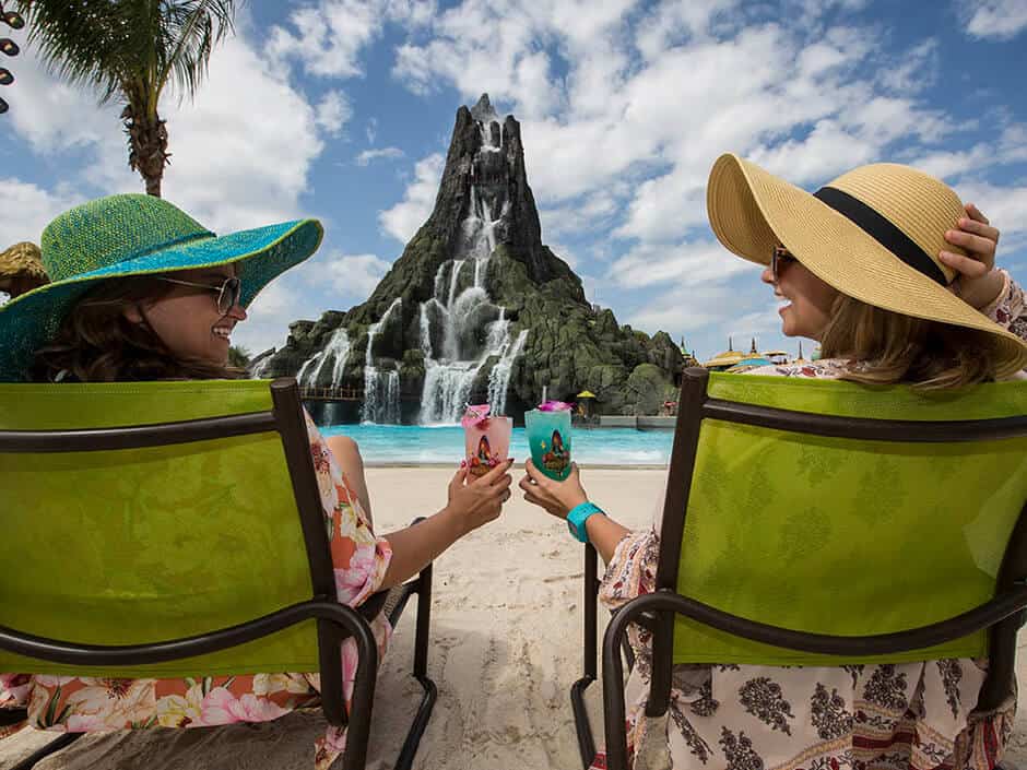 Two women in wide-brimmed hats sit in green chairs on a sandy beach, toasting with colorful drinks. Behind them, a tall rocky structure features cascading waterfalls under a partly cloudy sky. The tropical scenery creates a universally romantic Valentine's Day ambiance.