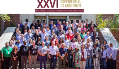 Large group of participants standing on outdoor steps beneath a sign for the 26th Experiential Classroom, an intensive entrepreneurship education professional development program.