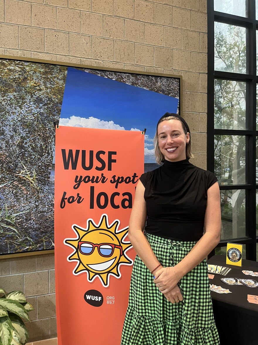 A women with short blond hair wearing a black top tucked into a green and black checkered skirt is standing in front of a sign that reads, "WUSF your spot of local."