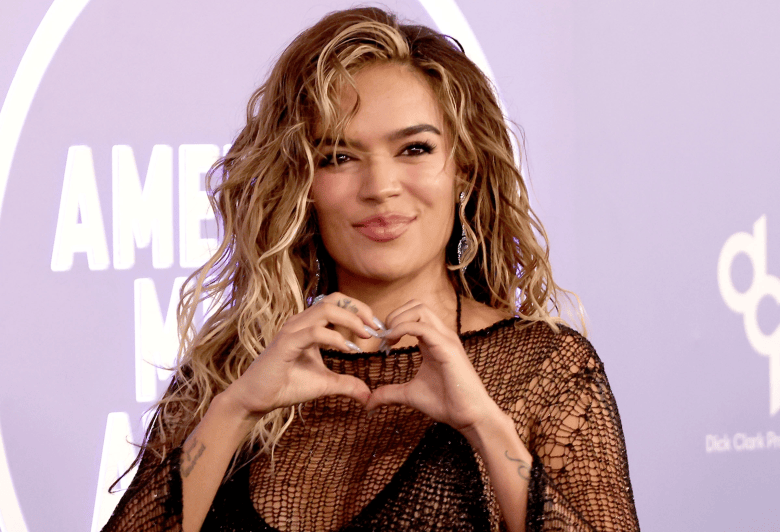 A red carpet portrait photograph by Kathy Hutchins showing Karol G at MGM Grand Garden Arena in Las Vegas, Nevada on May 25, 2026. Captured in a medium close-up, singer Karol G smiles at the camera while forming a heart shape with both hands over her chest. She wears a black mesh net top with long sleeves, drop earrings, and long wavy blonde hair with dark roots, standing in front of a purple event backdrop featuring white lettering under bright venue flash photography.
