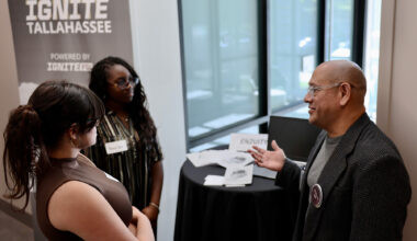 A man wearing glasses and a blazer speaks to two young women beside a table set up with informational materials near an "Ignite Tallahassee" banner.