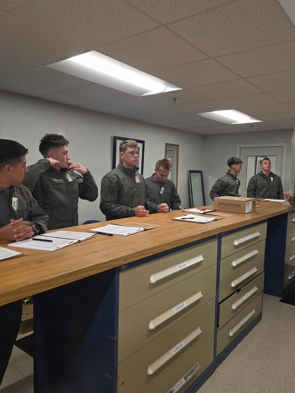 A group of young military personnel in olive/sage green flight suits are standing at a large wooden-topped counter with built-in drawers in what appears to be a military supply or equipment issue room. They appear to be processing paperwork, likely during an initial gear or equipment issue process. A box labeled 'McRae Footwear' is visible on the counter, suggesting this may be a uniform or equipment distribution event.