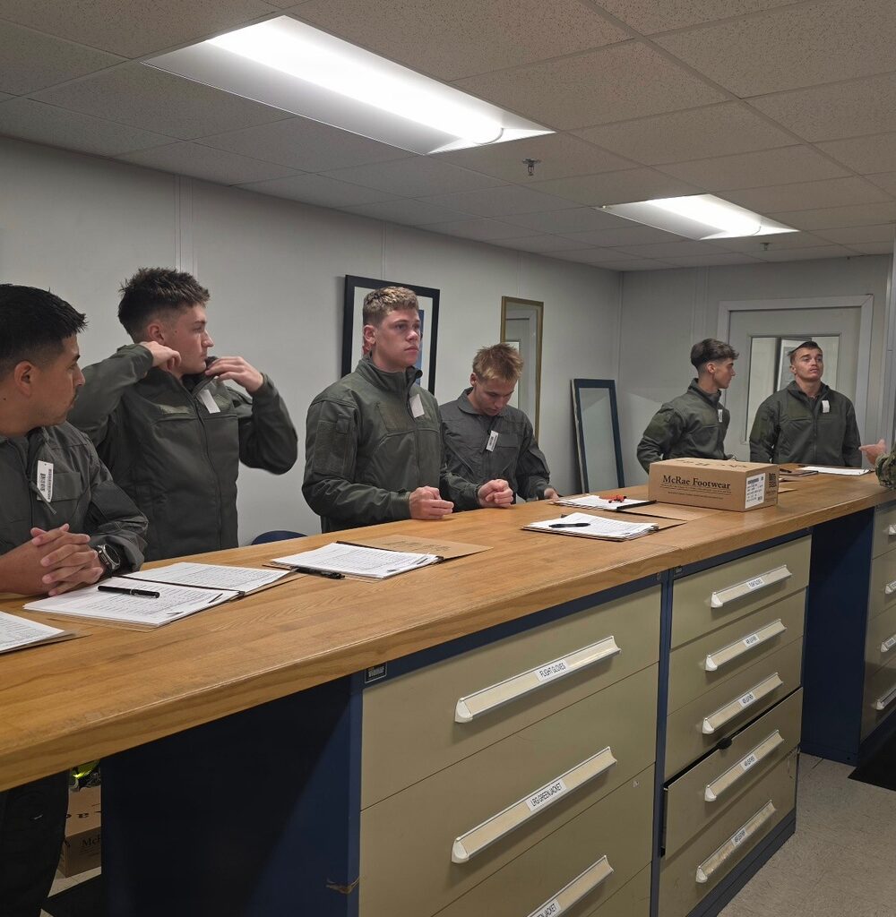 A group of young military personnel in olive/sage green flight suits are standing at a large wooden-topped counter with built-in drawers in what appears to be a military supply or equipment issue room. They appear to be processing paperwork, likely during an initial gear or equipment issue process. A box labeled
