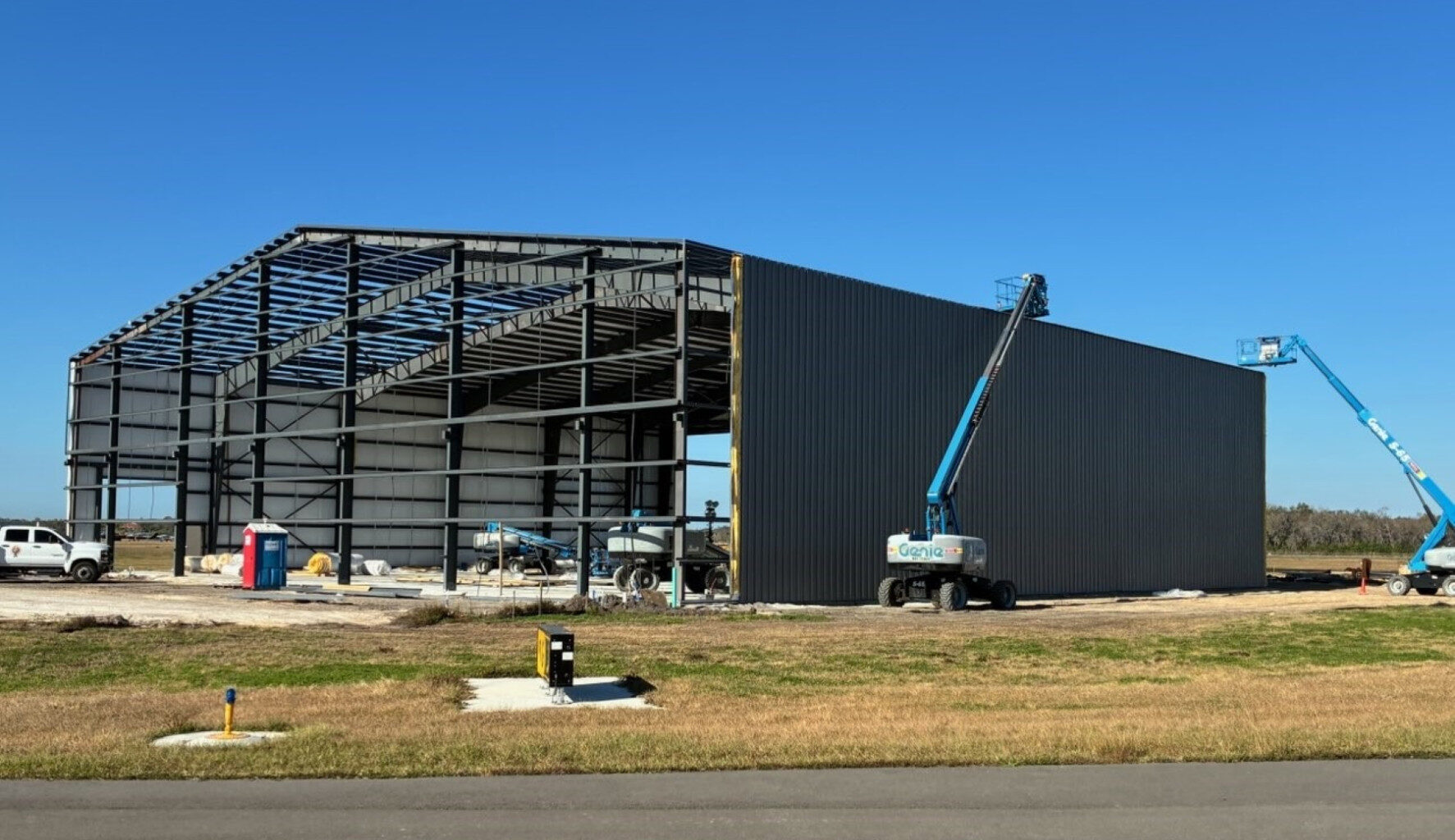 Large steel-frame aircraft hangar under construction, with cranes and lifts working on the exterior