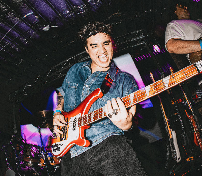 A concert performance action photograph by Dave Decker showing Andres Hernandez of Rohna at Crowbar in Ybor City, Florida on May 15, 2026. Captured in a dynamic low-angle shot on a dark stage, bass player Andres Hernandez plays an orange-and-white Rickenbacker-style electric bass while leaning back with an expressive open-mouthed expression. He wears a dark headband, short-sleeved blue denim button-up shirt, and dark trousers under bright concert stage flash lighting, with stage trussing and another band member partially visible in the background.