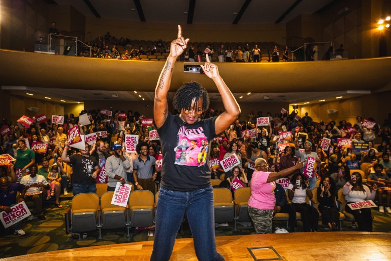 A high-energy rally photograph by Dave Decker showing Florida Rep. Angie Nixon at University of South Florida in Tampa, Florida on Oct, 4, 2026. Captured from a low-angle perspective on stage facing the audience, Representative Angie Nixon stands center stage celebrating with both arms raised pointing upward to the ceiling. She wears glasses, short dreadlocks, a dark t-shirt with a colorful graphic print, and blue jeans, backed by a cheering auditorium crowd holding pink and white campaign signs beneath upper balcony seating.